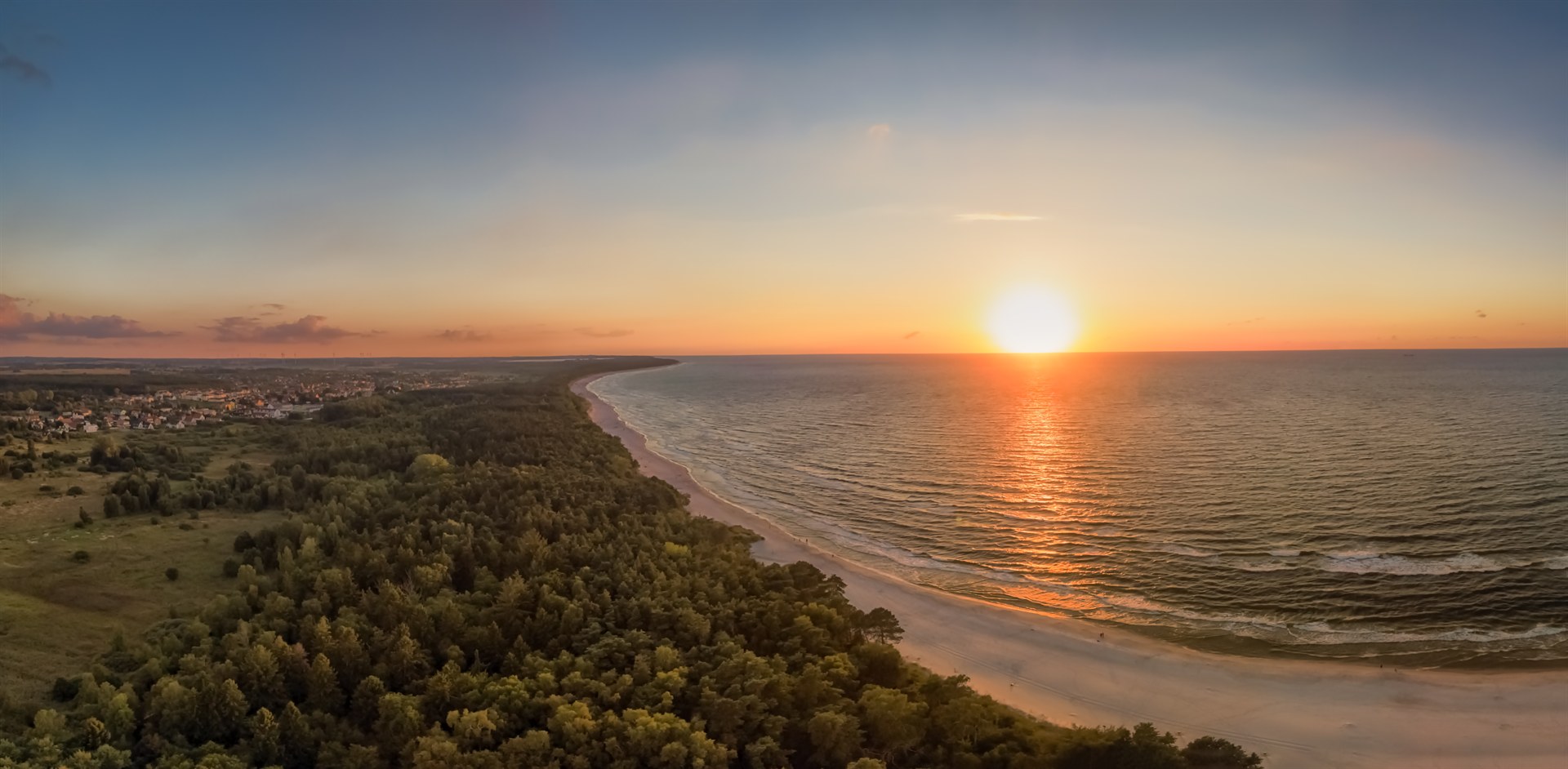 Zachód słońca nad morzem z odbiciem na wodzie, piaszczysta plaża i pas gęstego lasu oraz wiejska zabudowa