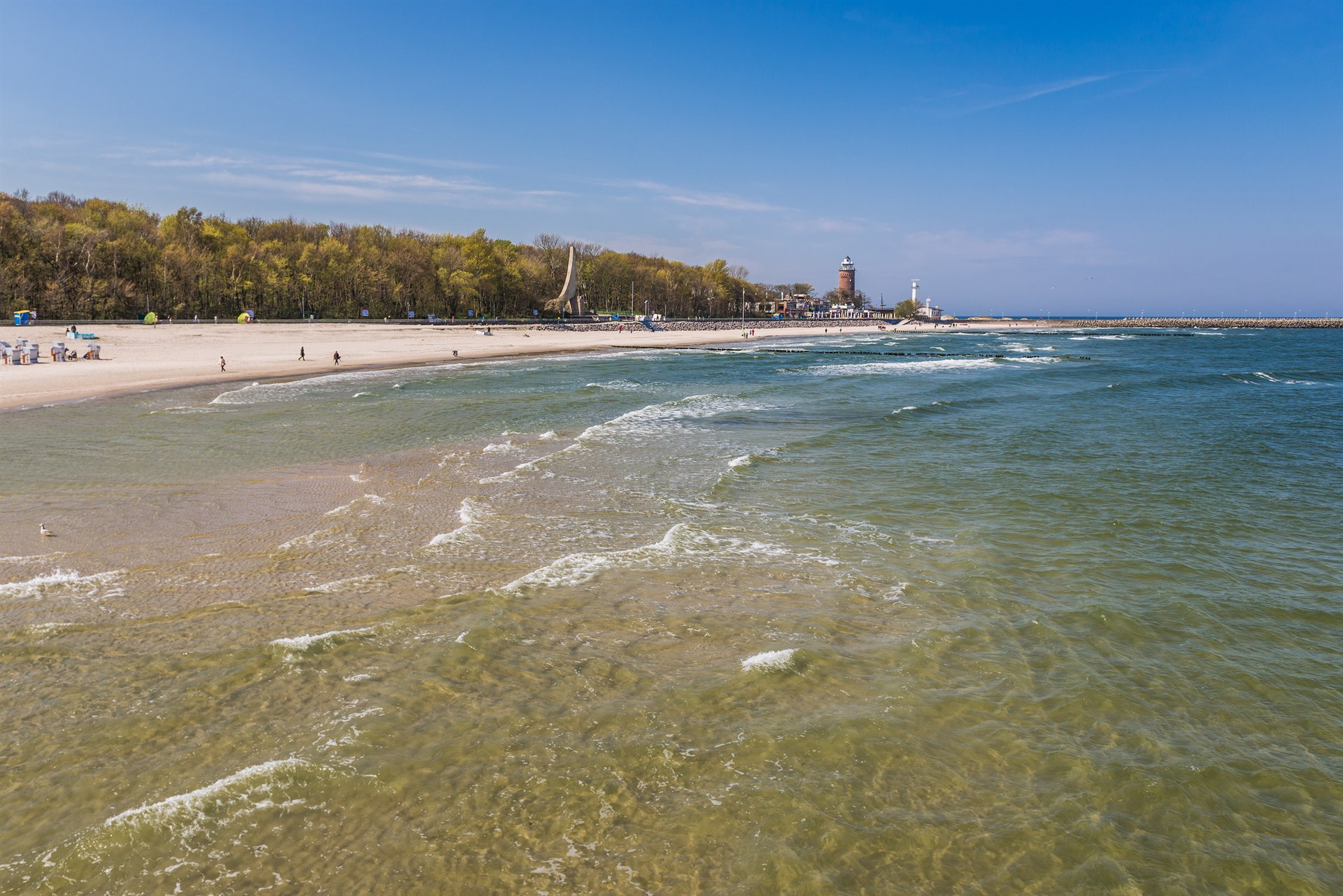 Piaszczysta plaża z niewielkimi falami na zielonkawej wodzie, lasem w tle oraz latarnią