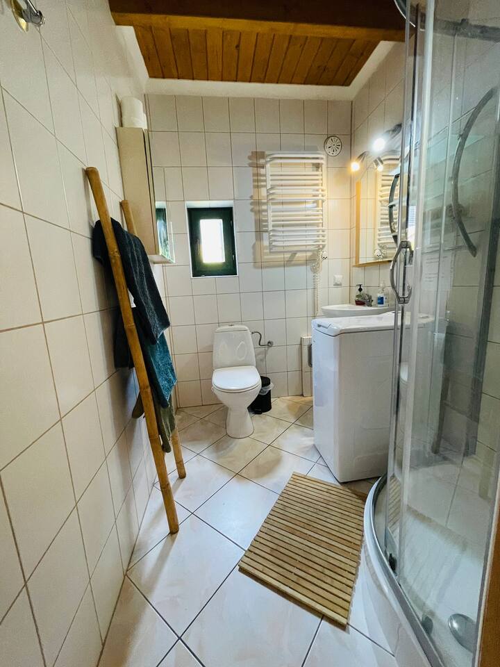Łazienka z białymi płytkami, narożną kabiną prysznicową, toaletą, pralką, drewnianą drabinką z ręcznikami i sufitowymi deskami