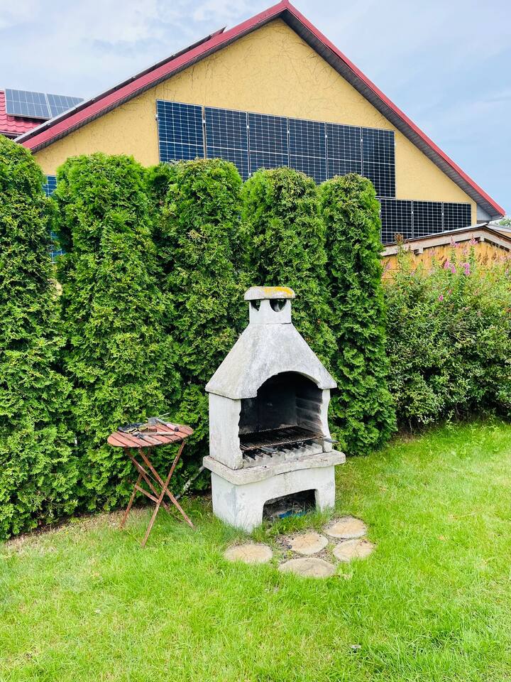 Betonowy grill ogrodowy z rusztem przed wysokim zielonym żywopłotem, obok mały składany stolik