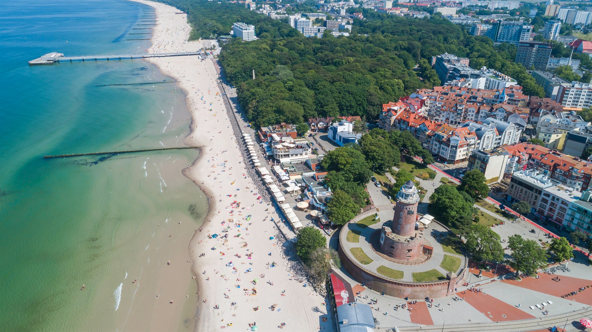 Widok z lotu ptaka na szeroką piaszczystą plażę z molo, czerwonym ceglastym latarniowcem i nadmorską promenadą